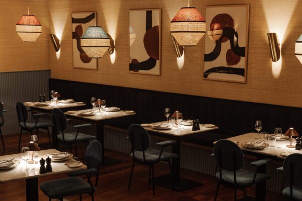 A warmly lit, modern restaurant with tables set for dining, abstract art on beige walls, woven pendant lights, upholstered chairs, and wooden flooring, creating an inviting and stylish ambience.