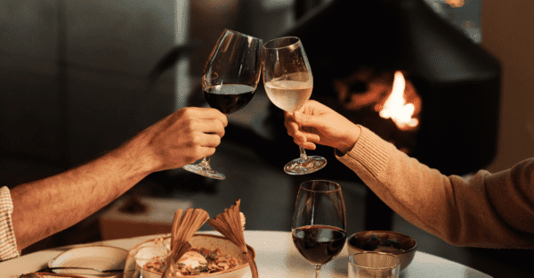 Two people clink wine glasses—one filled with red wine, the other with white—over a dining table, with food and a burning fireplace visible in the warm, cosy background.