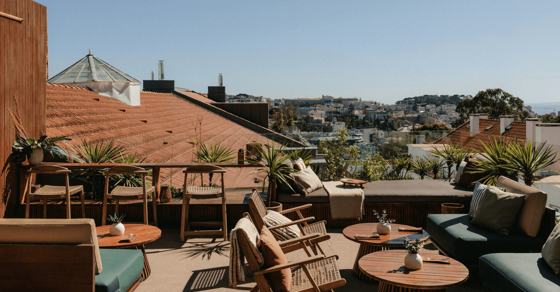 A rooftop terrace with wooden furniture, potted plants, and views of red-tiled roofs and a distant cityscape under a clear blue sky.