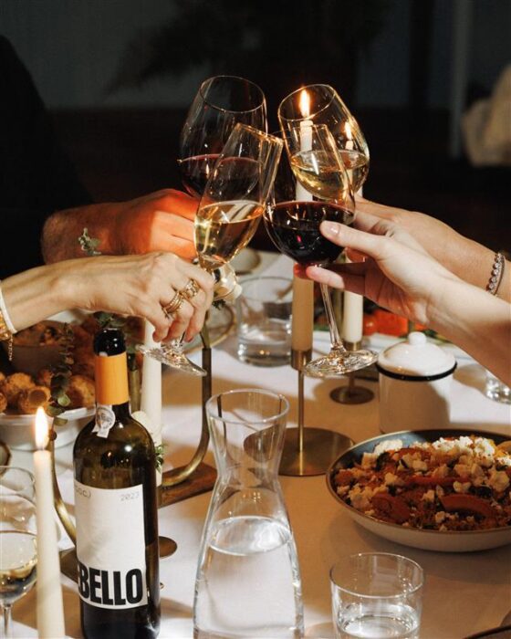 Four people clink wine glasses over a candlelit dinner table set with food, a bottle of wine labelled 