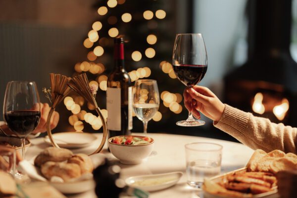 A hand holds a glass of red wine over a festive dinner table set with bread, salad, wine, and water. Warm lights and a blurred Christmas tree create a cosy, celebratory atmosphere in the background.