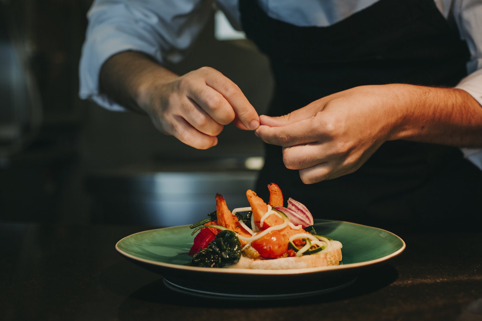 A chef in a white shirt and dark apron carefully garnishes a dish of prawns, vegetables, and greens arranged on a green-rimmed plate, focussing on presentation in a dimly lit kitchen.