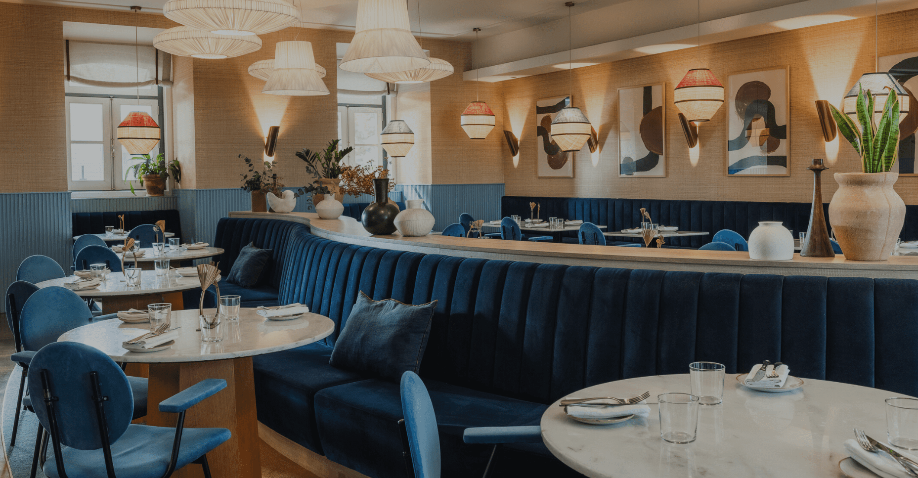 A modern restaurant interior with blue velvet seating, marble tables set with glassware, and decorative pendant lights. Neutral walls feature abstract art whilst large vases with plants accent Blue Bistrot for Lisbon Restaurant Week.