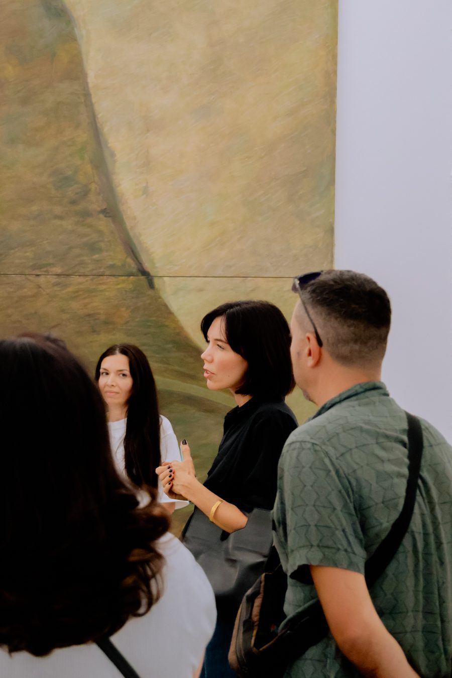 A group of people stands indoors, attentively listening to a woman speaking in front of a large abstract painting with earth-toned colours.