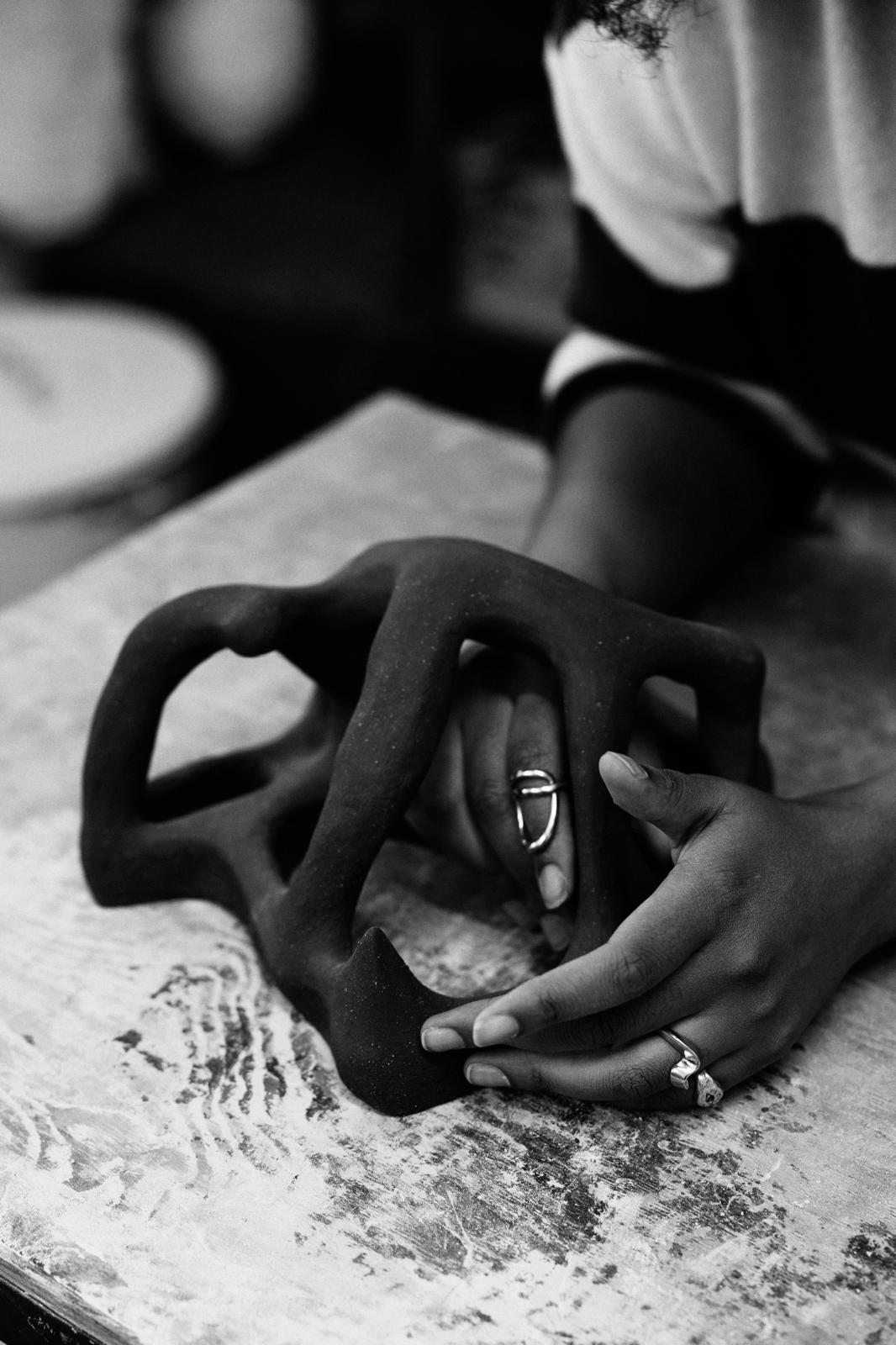 A person shapes a dark, abstract ceramic sculpture on a textured table. Their hands, adorned with rings, gently hold the piece. The image is in black and white.