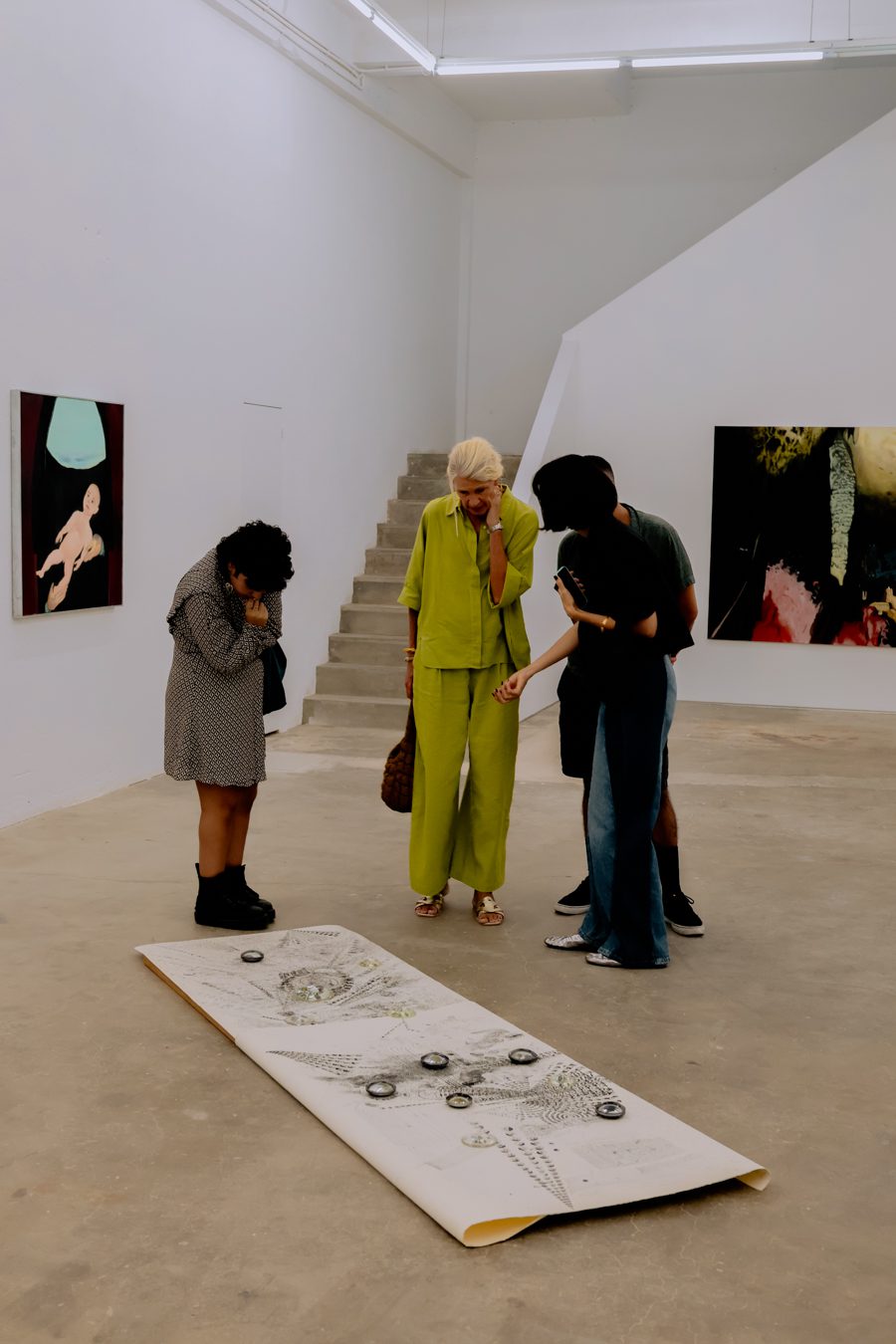 Four people stand in a modern art gallery, closely examining a large artwork placed on the floor; paintings hang on white walls in the background.