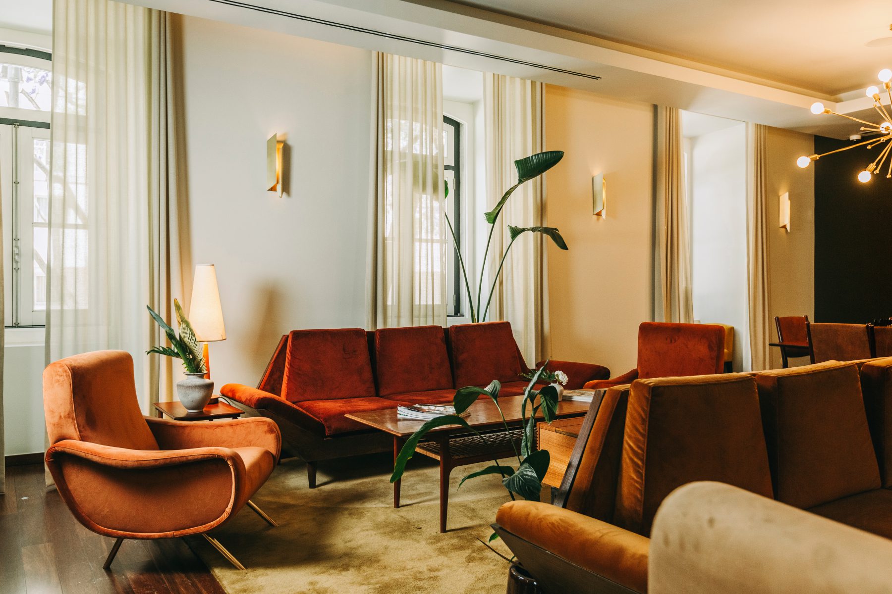 A stylish living room with mid-century modern furniture, including orange armchairs and a sofa, a wooden coffee table, potted plants, and large windows with sheer curtains allowing natural light inside.