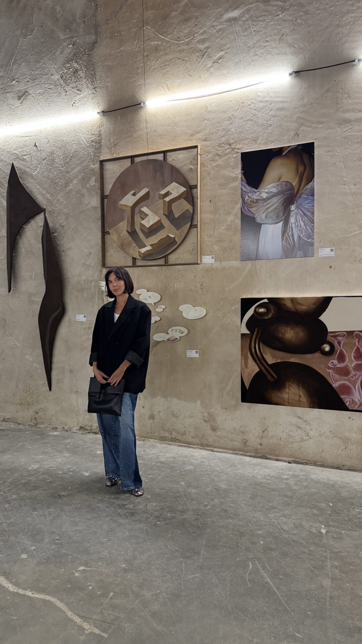 A woman with shoulder-length dark hair, wearing a black blazer, jeans, and black shoes, stands in front of abstract art pieces on a textured beige wall in a gallery space with concrete floors.