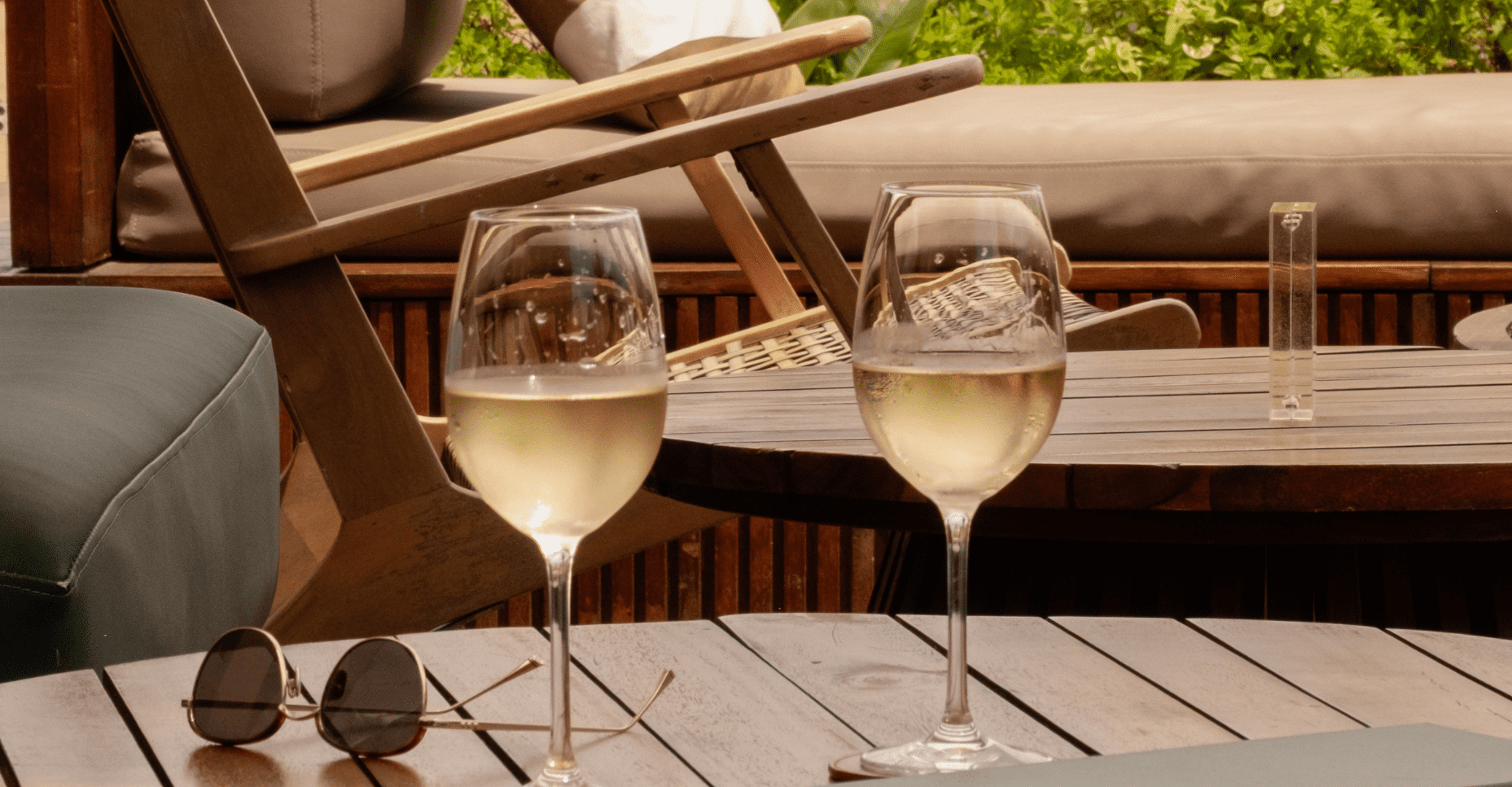 Two glasses of white wine sit on a round wooden outdoor table beside a pair of sunglasses, with loungers and greenery in the background, suggesting a relaxed, sunny setting.