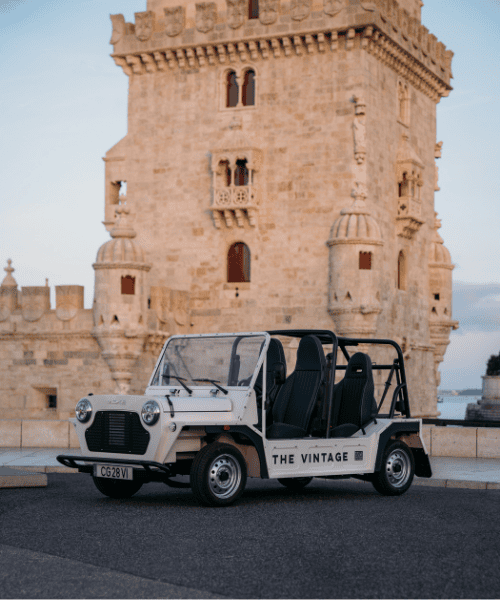 A white open-top vintage car labelled "THE VINTAGE" is parked on a street in front of a historic stone tower with ornate carvings and arched windows.