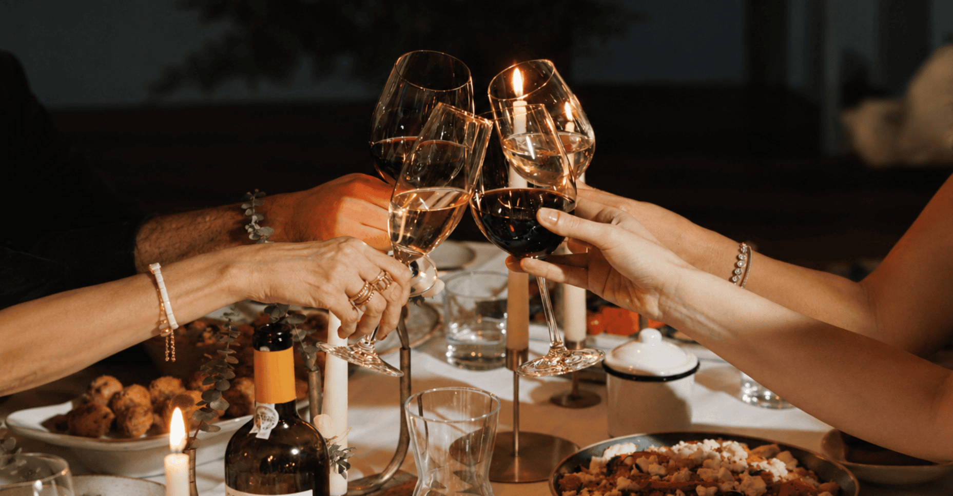 Four hands clink wine glasses over a candlelit dinner table set with food, candles, and a wine bottle, creating a festive and celebratory atmosphere.