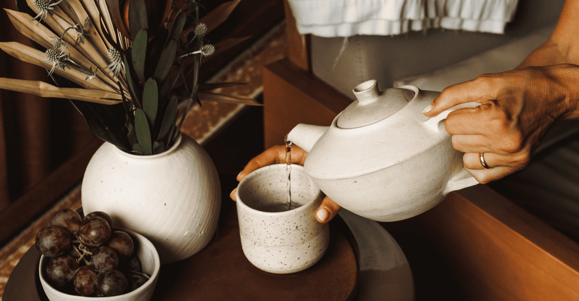 A person pours hot water from a ceramic teapot into a matching mug, next to a bowl of grapes and a vase of dried plants.