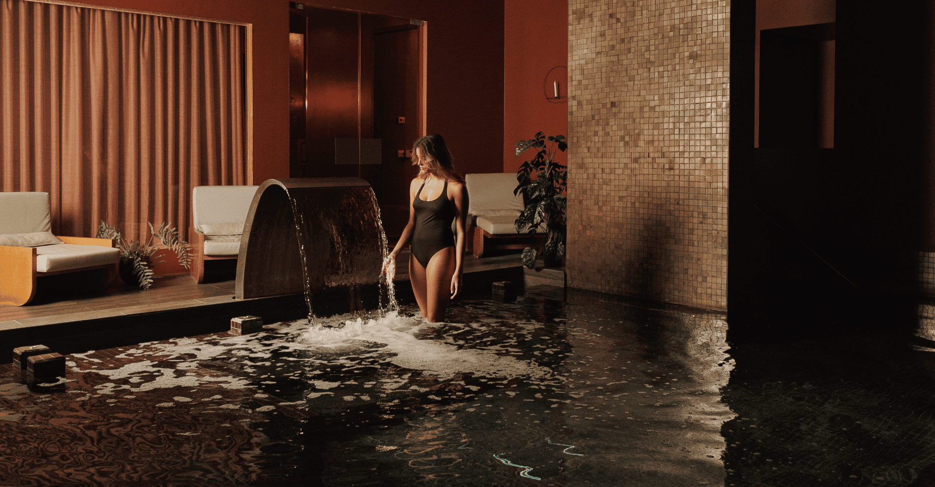 A woman in a black swimming costume stands in a spa pool under a water feature.